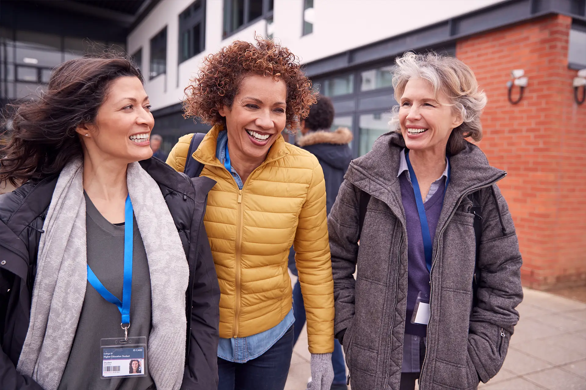 Three women with badges chat and smile outside a modern Leeds building, possibly volunteering for a local charity.