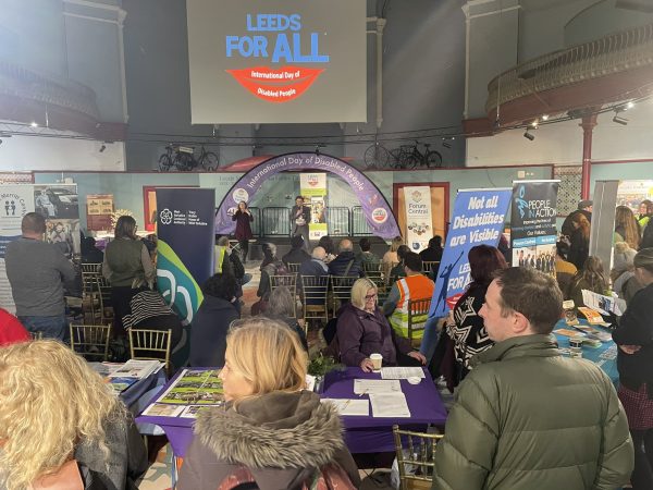 People at a disability awareness event in Leeds with info stalls, banners and a stage talk, by Doing Good Leeds.