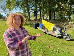 Volunteer with curly hair gestures to a yellow-sailed homemade vehicle at an off-grid project in Armley, Leeds, for Doing Good Leeds.