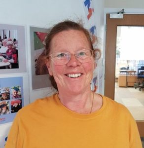 Smiling woman celebrates 25 years volunteering at MAE Care, Leeds, standing by a photo wall indoors.