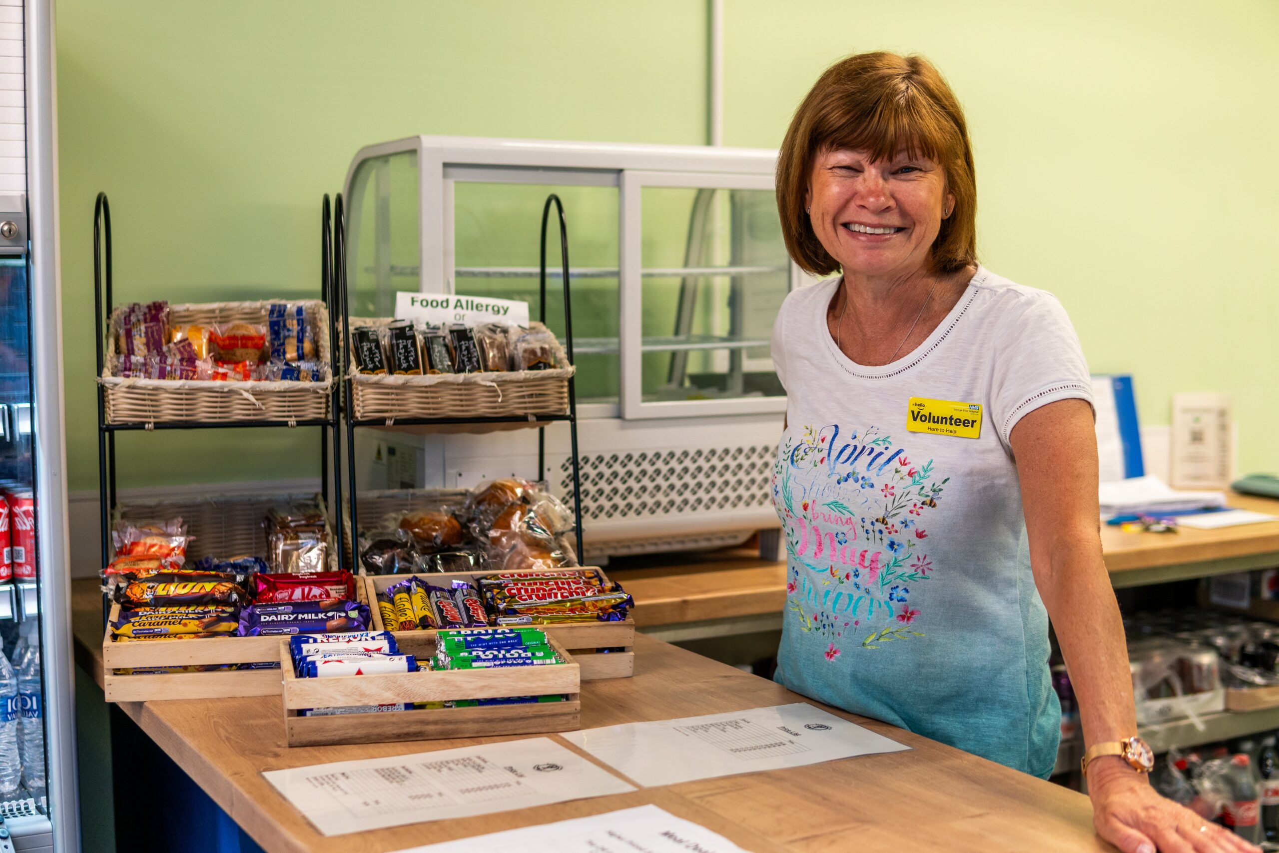 Volunteer with short brown hair welcomes visitors behind a snack counter in a bright Leeds café, supporting local charity work.
