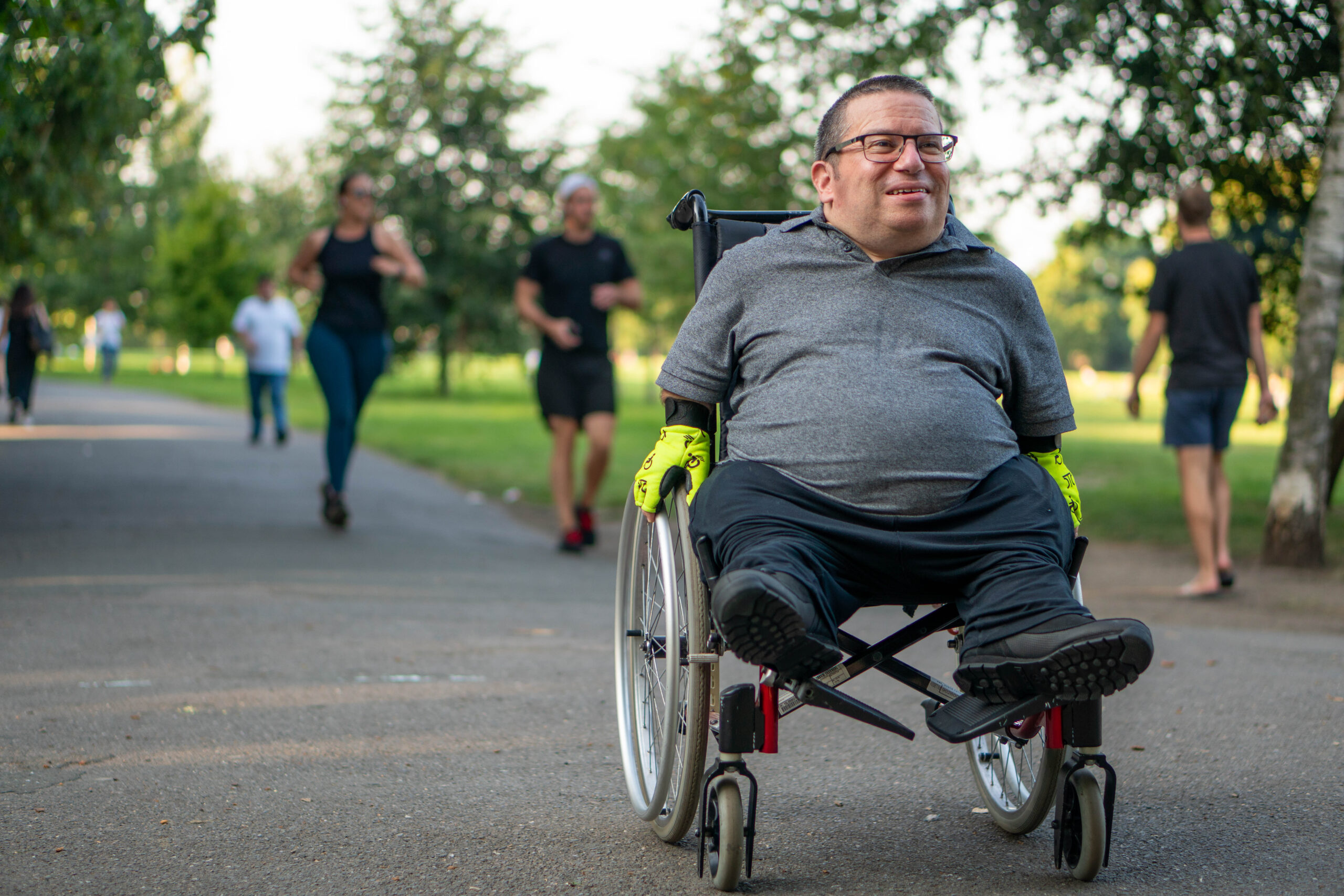 Man in wheelchair wearing yellow gloves moves along leafy park path, with others walking nearby, Leeds community volunteering.
