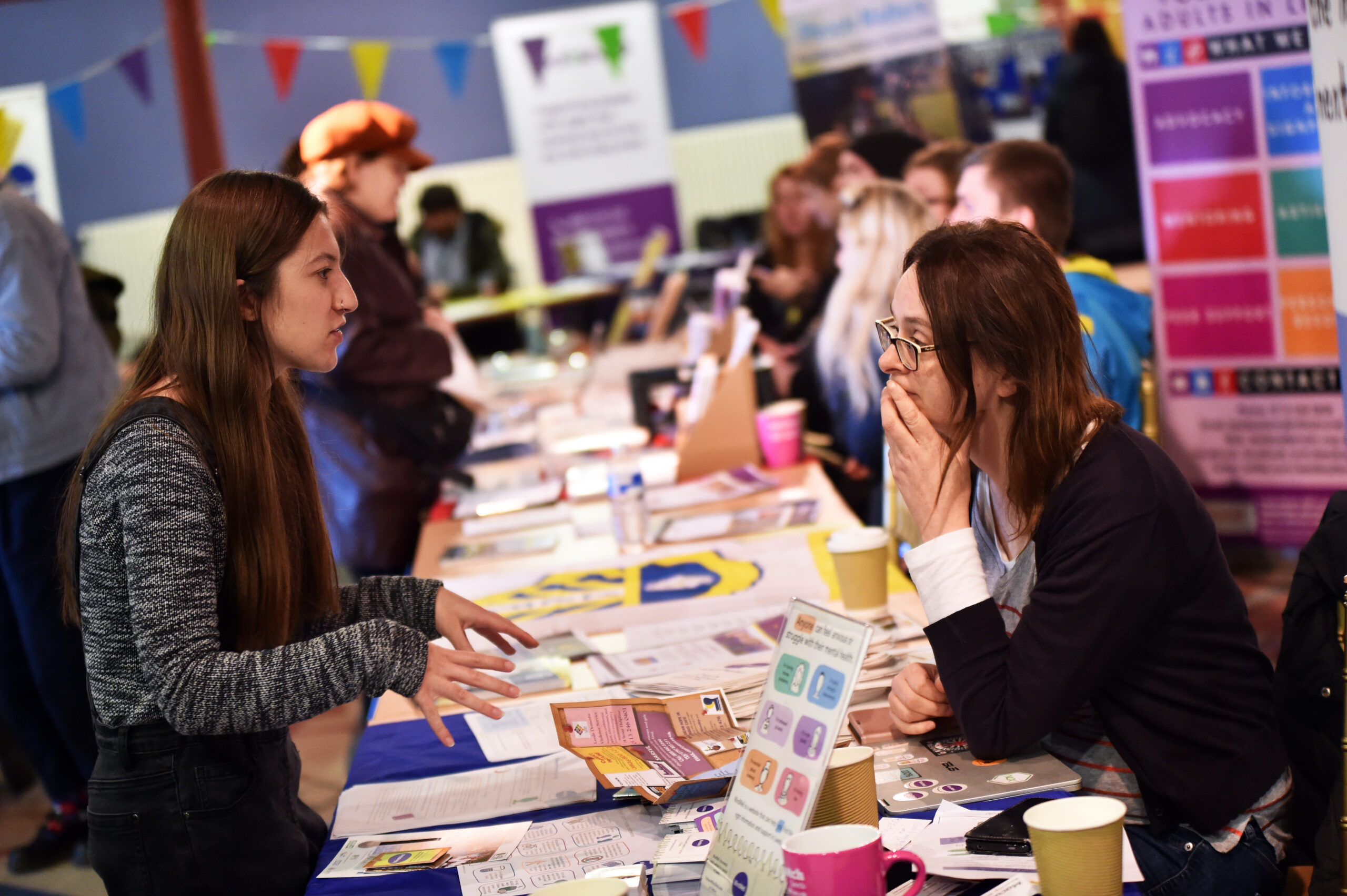 Two women chat at a busy Leeds volunteering fair, sharing info by a table with leaflets as others talk nearby.